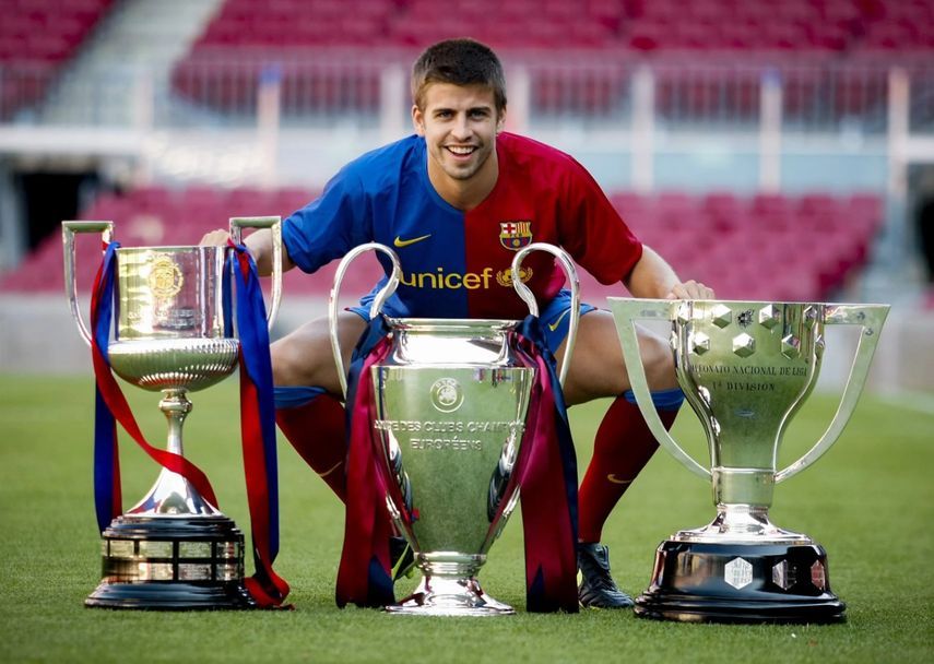 صورة تذكارية لمدافع برشلونة جيرارد بيكيه مع كؤؤس دوري أبطال أوروبا والدوري الإسباني وكأس ملك إسبانيا